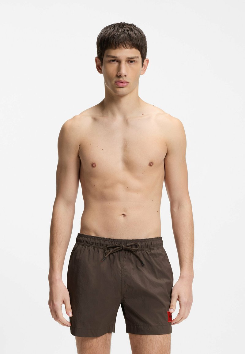Jeune homme debout torse nu portant un short de bain marron foncé avec cordon de serrage sur fond blanc.