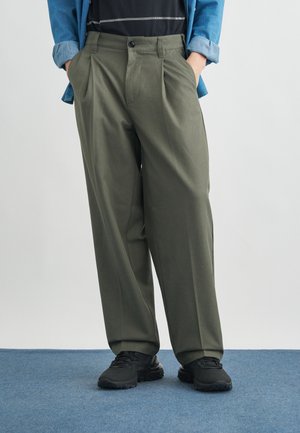 Άτομο που στέκεται με τα χέρια στις τσέπες, φοράει χαλαρά ελαιχρώματα παντελόνια, μαύρα αθλητικά παπούτσια, σκούρο κορυφαίο ρούχο και μπλε μακρυμάνικο πουκάμισο σε μπλε χαλί.