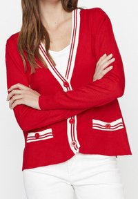 Mujer que lleva un cárdigan rojo con ribete blanco, abotonado al frente y con dos bolsillos delanteros, combinado con pantalones blancos y con los brazos cruzados.