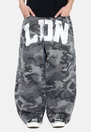 Persona che indossa pantaloni larghi mimetici grigi con grandi lettere bianche "LDN" sul davanti, mani nelle tasche e scarpe da ginnastica bianche e nere.