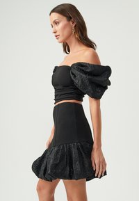 Čierny dvojdielny outfit obsahujúci priliehavý skrátený top s nafúknutými rukávmi a rozšírenú sukňu s textúrovaným volánom na okraji.