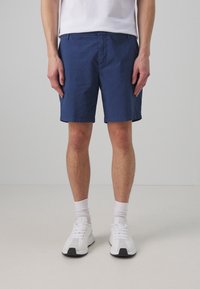 Homme portant un short bleu marine, des chaussettes blanches, des baskets blanches et un t-shirt blanc rentré, se tenant contre un fond gris clair uni.