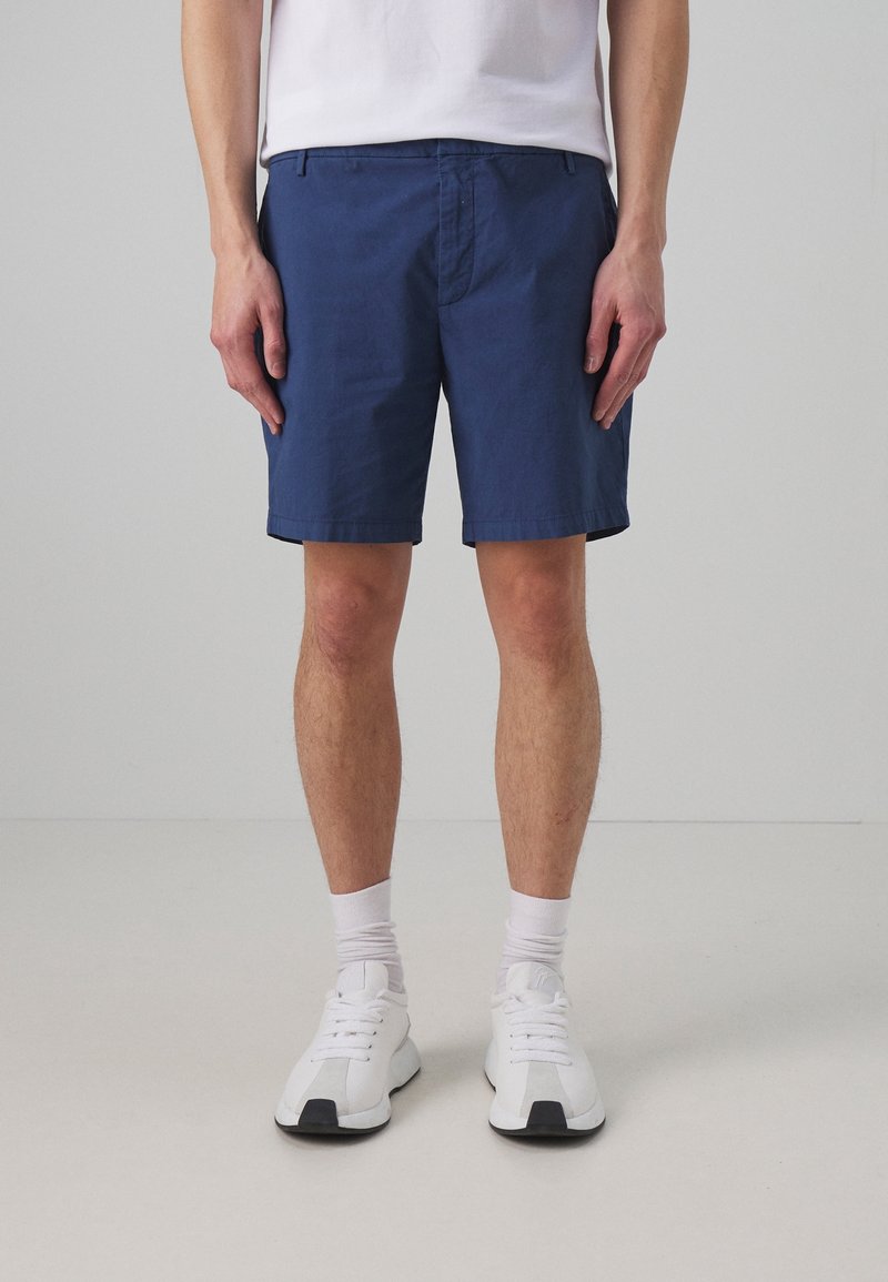 Homme portant un short bleu marine, des chaussettes blanches, des baskets blanches et un t-shirt blanc rentré, se tenant contre un fond gris clair uni.