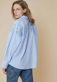 Camicia azzurro chiaro con un design ricamato sul retro, maniche lunghe e polsini con bottoni, abbinata a jeans blu con pattern.