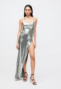 Zilveren jurk met pailletten, aansluitende pasvorm, zijsplit en verstelbare bandjes, met een gladde, glanzende textuur. Zwarte sandalen met hak maken de outfit compleet.