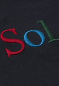 Izvezeni tekst koji glasi "Sol" na tamnoj pozadini od tkanine. Slova su u crvenoj, plavoj i zelenoj niti, s mješavinom podebljanih i razigranih oblika.