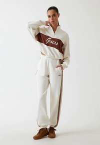 Valge lukuga dressipluus pruunide triipude ja "Guess" logoga. Sobivad valged dressipüksid pruunide külgtriipude ja taskutega. Pruunid tossud.