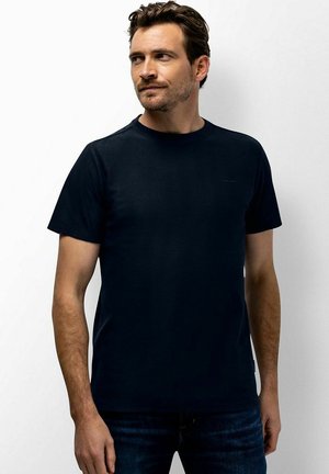 Man draagt een effen donkerblauw T-shirt en donkere spijkerbroek, kijkt naar links tegen een witte achtergrond.
