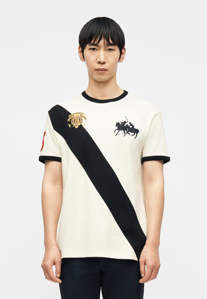 Νέος άνδρας που φοράει λευκό πόλο με μαύρη διαγώ νια ρίγα, λογότυπο παίκτη πόλο στο στήθος και κόκκινο αριθμό 3 στο μανίκι, στέκεται μπροστά σε λευκό φόντο.