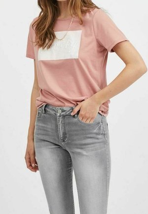 Jasnoróżowa koszulka z białym prostokątnym nadrukiem z napisem "Lovely" w stylu kursywy, zestawiona z dopasowanymi szarymi dżinsami. Miękki bawełniany materiał.