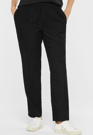 Person wearing black corduroy pants and white sneakers, standing with hands in pockets against a plain background.