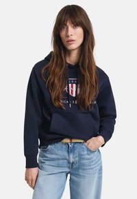 Donkerblauwe hoodie met een voorkraag en een logo met rode en witte strepen. Gecombineerd met lichtgekleurde spijkerbroek en een gebroken riem.