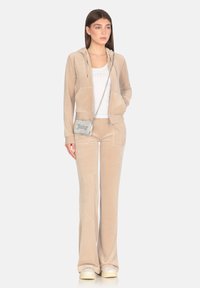 Tuta in velluto beige con cappuccio zip, pantaloni a gamba larga e borsa a tracolla argento con testo. Texture liscia e vestibilità rilassata.