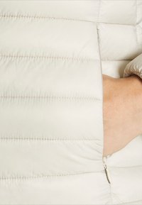 Svetlo béžová péřová bunda s vertikálnym prešívaním, zipsovými vreckami a detailmi na elastických manžetách. Hladká textúra, ľahký materiál.