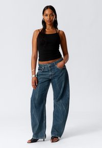 Weit geschnittene, dunkelblaue Jeans aus Denim mit einem ausgewaschenen Finish, getragen mit einem enganliegenden schwarzen Tanktop und schwarzen Absatzsandalen, betont durch silbernen Schmuck.