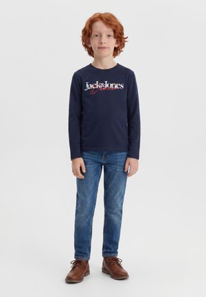 Rudowłosy chłopiec stoi, ubrany w granatową koszulkę z długim rękawem z napisem „Jack & Jones”, niebieskie dżinsy i brązowe buty sznurowane na białym tle.