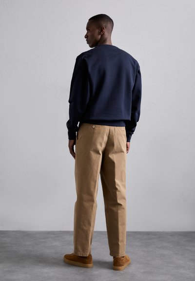 Navy blue sweatshirt with long sleeves, paired with light brown, relaxed-fit trousers. Model wearing brown suede shoes. Simple, minimalist design.