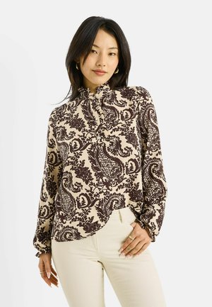 Mujer con cabello largo y negro, vestida con una blusa beige con estampado floral oscuro de estilo cachemira y pantalones crema, de pie con una mano en la cadera.