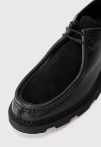 Schwarzer Lederschuh mit einem weichen schwarzen Wildledereinsatz, dicker schwarzer Gummisohle und detaillierter Naht entlang der Kante.