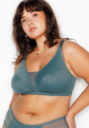 Donna con capelli castani di media lunghezza che indossa un reggiseno verde acqua testurizzato e biancheria intima abbinata, in piedi con la mano sul fianco su sfondo bianco.