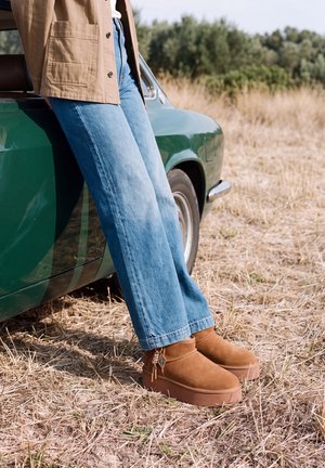 Une personne en jean bleu et en bottes marron clair s’appuie contre une voiture verte dans un champ d’herbe sèche.