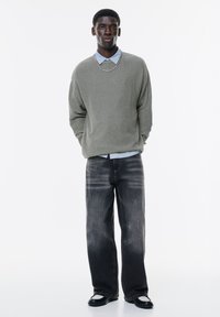 Grå ribbet sweater over en lyseblå skjorte med krave. Bæres med slidte, løse sorte jeans og sorte sko med hvide detaljer. Stående stilling.
