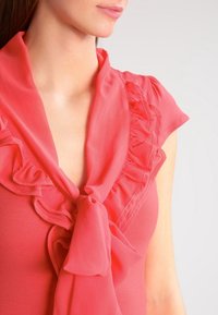 Coral blouse with a ruffled neckline and bow detail, made of lightweight fabric. Short sleeves, smooth texture, and subtle sheen.