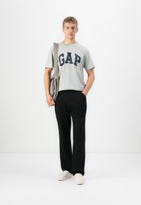 Grå kortärmad t-shirt med "GAP" i marinblått, svarta byxor och ljusa skor. En grå budväska hängande över ena axeln.
