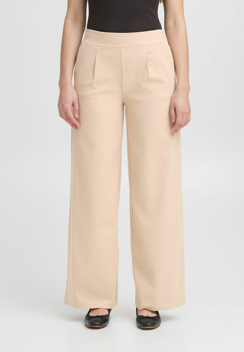 Pantaloni a gamba larga in tessuto beige chiaro. Presentano un'alta vita, pieghe davanti e una superficie liscia. Abbinati a ballerine nere.