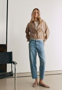 Donna in piedi al chiuso che indossa una giacca beige, una maglietta bianca, jeans azzurri e ballerine taupe accanto a una sedia nera.