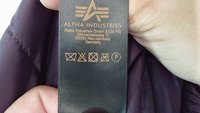 Zwarte verzorgingslabel van Alpha Industries, met wasymbolen en tekst in goud, bevestigd aan een donkerpaarse stof.