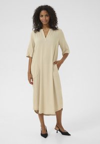 Robe midi beige avec un décolleté en V profond, des manches trois-quarts et deux grandes poches latérales. Texture lisse, silhouette fluide et coupe décontractée.