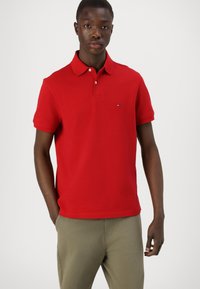 Polo rojo de algodón con cuello clásico, mangas cortas y un pequeño logotipo en el pecho. Combinado con chinos de color claro.