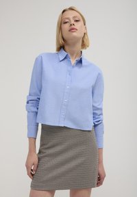 Giovane donna con capelli biondi corti che indossa una camicia blu chiaro con bottoni e una gonna a quadri marrone su uno sfondo semplice.