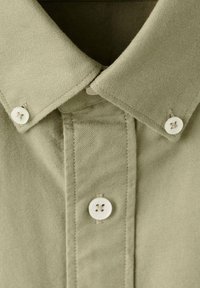 Camisa con cuello en tejido de algodón verde oliva claro que presenta botones de plástico blancos y detalles de costuras visibles en el cuello y la tapeta.