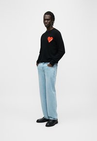 Pull noir avec un motif de cœur rouge indiquant "FUTURE", associé à un jean large bleu clair et des chaussures noires, posé devant un fond blanc.