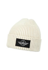 White knitted beanie with a ribbed texture. Features a black fabric patch with a circular emblem and star graphic with wing accents.