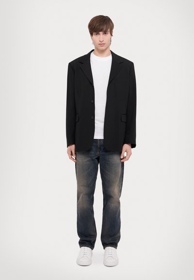 Schwarzer oversized Blazer mit zwei Knöpfen, Reverskragen und einer Brusttasche, getragen über einem weißen T-Shirt und kombiniert mit blauen Jeans und weißen Sneakers.