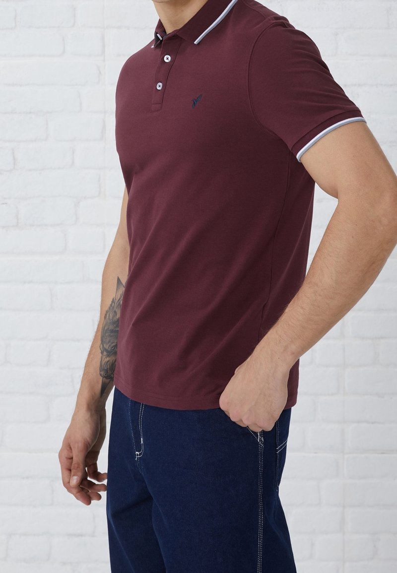 Homme portant un polo bordeaux avec des bordures blanches aux manches et un jean bleu foncé, debout devant un mur de briques blanches.