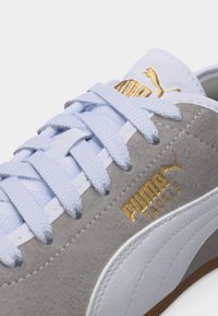 Κοντινό πλάνο γκρι και λευκού αθλητικού παπουτσιού Puma Tackle με ανοιχτό μπλε κορδόνια και χρυσά λογότυπα Puma στη γλώσσα και στο πλάι.