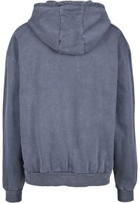 Kapucnis pulcsi tompa szürke színben, laza szabással, bordázott mandzsettákkal és heméssel, valamint puha anyagtextúrával. A hátoldalon varrási részletek láthatók.