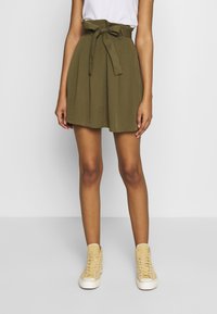 Olivgrüner Rock mit hoher Taille und Schleife, mit Falten und geradem Saum. Kombiniert mit beigen High-Top Sneakern.