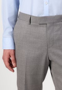 Pantaloni grigi su misura con una sottile texture, davanti piatto e tasche laterali, abbinati a una camicia azzurra a maniche lunghe.