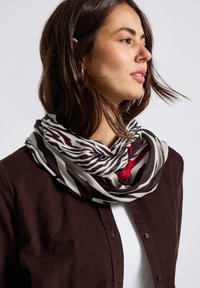 Una donna con capelli castani di lunghezza media indossa una sciarpa a zig-zag nera e bianca e una camicia marrone scuro con bottoni sopra una maglietta bianca.