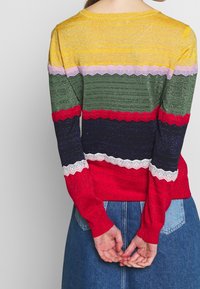 Maglione lavorato a maglia con strisce orizzontali in giallo, rosa, verde, rosso, blu navy e crema, con bordi ondulati; colori vivaci e texture interessanti.