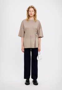 T-shirt oversize beige con un dettaglio a cerchio, abbinato a pantaloni larghi blu scuro. Il modello tiene una piccola borsa bianca e indossa scarpe nere.
