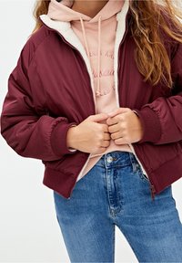 Bordeaux bomberjakke med ribbede manchetter, foret med beigefarvet fleece, båret over en lyserød hættebluse og blå jeans. Lynlåslukning foran.
