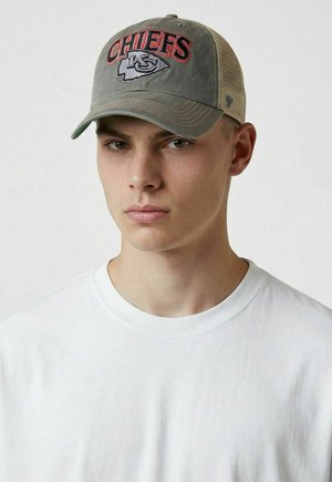 Junger Mann trägt eine grau-beige Kansas City Chiefs Cap und ein schlichtes weißes T-Shirt mit Rundhalsausschnitt, schaut geradeaus mit neutralem Ausdruck.