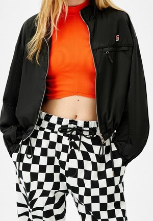 Personne portant une veste noire zippée, un top court côtelé orange vif et un pantalon à cordon à carreaux noir et blanc.