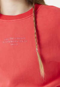 Sudadera de color coral con un cuello redondo de canalé y texto bordado "HAZ EL VIAJE A LUGARES DESCONOCIDOS" en blanco, con una trenza larga cerca.